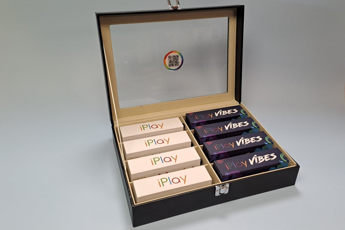 An open black presentation box with a glass lid, containing five beige iPlay boxes and four dark purple VIBES boxes, set against a light gray background.